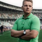Man in green polo stands confidently with arms crossed in a sports stadium.
